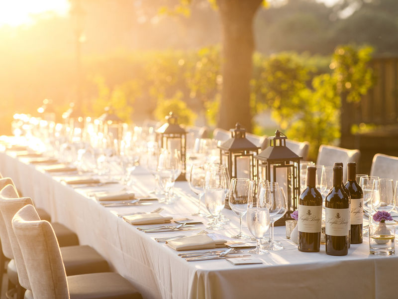 Outdoor dining table set for a meal, with wine bottles, glassware and lanterns in the evening sunlight.